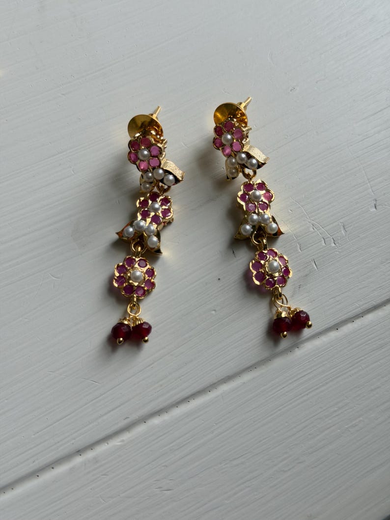 May include: A pair of gold-toned earrings with a floral design. Each earring features pink and white flower-shaped accents, small white pearls, and dangling red beads. The earrings are displayed on a white surface.