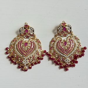 Può includere: Un paio di orecchini dorati con un design a forma di cuore. Gli orecchini presentano pietre preziose rosse, perle bianche e perline rosse pendenti. L'intricato design è posto su uno sfondo bianco.