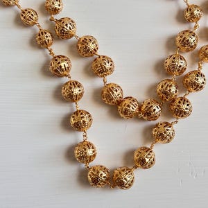 Può includere: Primo piano di due collane dorate con intricate perline sferiche traforate. Le perline sono infilate con delicate catene d'oro. Le collane sono esposte su una superficie bianca, mostrando il loro design dettagliato e il colore caldo.