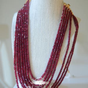 May include: A multi-strand necklace made of deep red faceted beads. The necklace is displayed on a white mannequin bust. The necklace is made by Maharani Jewelz, which is written on the bust.