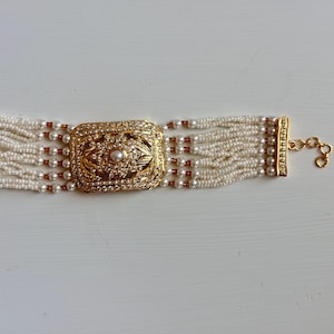 May include: A gold choker necklace featuring multiple strands of small, white beads. The central element is a rectangular gold piece adorned with clear rhinestones and a central pearl. The necklace is finished with a gold clasp.