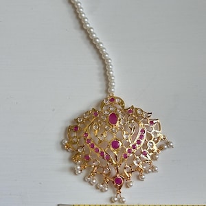 Może przedstawiać: Złoty nakrycie głowy z kwiatowym wzorem, z czerwonymi i przezroczystymi kamieniami szlachetnymi. Nakrycie głowy jest połączone z pasmem małych białych pereł. Element ma szerokość około 5 cm.