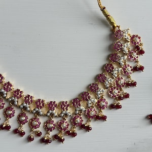 May include: A gold-toned necklace with a floral design. The necklace features small, round, red gemstones and white pearls set within the floral elements. Small red beads dangle from the bottom of each floral cluster.