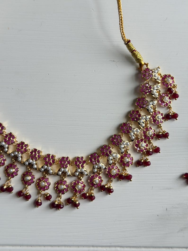 May include: A gold-toned necklace with a floral design. The necklace features small, round, red gemstones and white pearls set within the floral elements. Small red beads dangle from the bottom of each floral cluster.