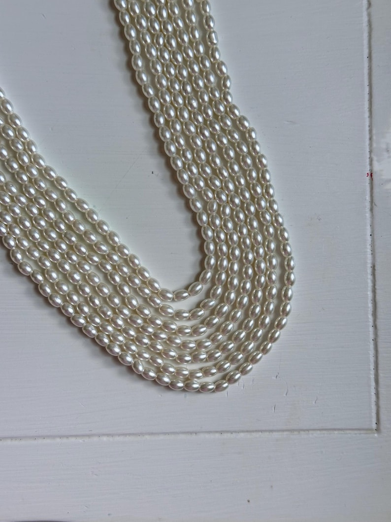 Op de afbeelding: Een meerstrengs parelketting in een cr&egrave;mewitte kleur. De ketting heeft meerdere strengen ovaalvormige parels, waardoor een gelaagd effect ontstaat. De parels hebben een subtiele glans en de ketting wordt gepresenteerd op een wit oppervlak.