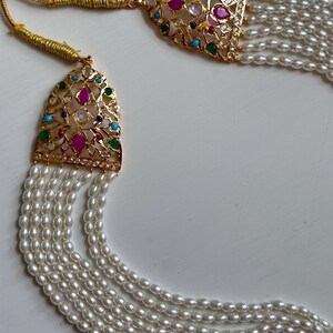 Op de afbeelding: Een goudkleurige ketting met meerdere strengen witte parels. De ketting heeft twee sierlijke hangers met kleurrijke edelstenen, waaronder roze, groene, blauwe en heldere stenen. De hangers hebben een filigraan ontwerp.