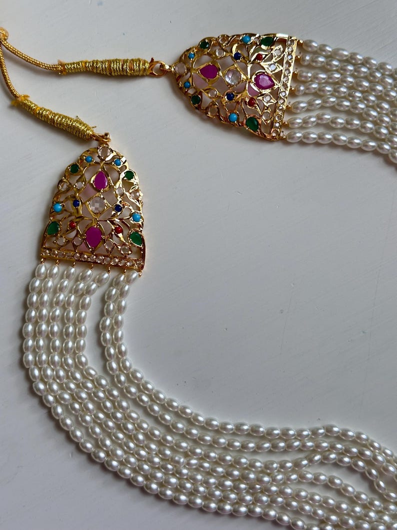Op de afbeelding: Een goudkleurige ketting met meerdere strengen witte parels. De ketting heeft twee sierlijke hangers met kleurrijke edelstenen, waaronder roze, groene, blauwe en heldere stenen. De hangers hebben een filigraan ontwerp.