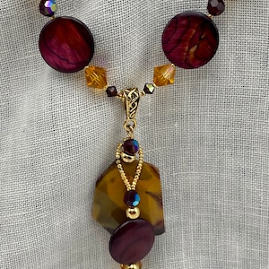 May include: A gold necklace with a brown and purple stone pendant. The pendant is made of multiple stones and has a gold chain attached to it. The necklace is made of gold beads and brown and purple stones.