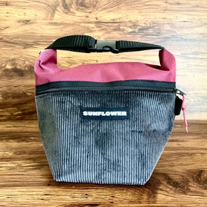 Op de afbeelding: Een zwarte corduroy lunchtas met een roze rits en een zwarte band. De tas heeft een zwart label met de tekst "SUNFLOWER".