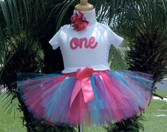 May include: A white t-shirt with the word "one" in pink script on the front, paired with a multi-colored tulle tutu with a pink satin bow. The tutu has layers of pink, blue, and purple tulle.
