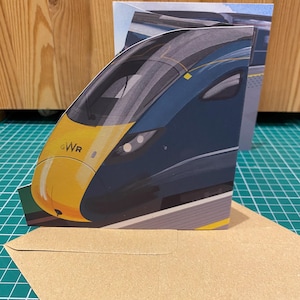 May include: A greeting card depicting a high-speed train, with a yellow and navy blue front. The card displays the letters "GWR". It is presented with a brown envelope, suitable for sending. The card is on a green surface.