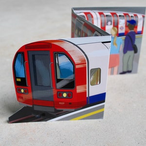 May include: A red and white London Underground train with a blue stripe. The train is on a track with a yellow line. There are people waiting on the platform.