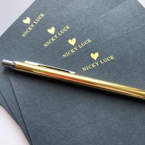 May include: Four dark grey note cards with gold foil hearts and the text "Nicky Luck" printed on each card. A gold pen is lying on top of the cards.