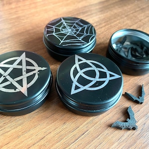 May include: Four black metal containers with different silver designs. The designs include a pentagram, a triquetra, and a spider web. There are also three small black bat-shaped objects.