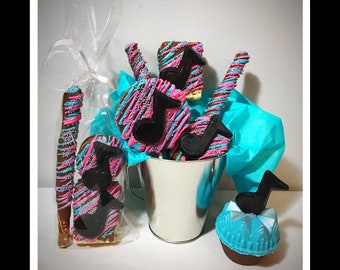 May include: A collection of chocolate-covered treats, including pretzels, cookies, and a cupcake, decorated with pink and blue drizzle and black musical notes. The treats are arranged in a silver bucket and cellophane wrap, with blue tissue paper.