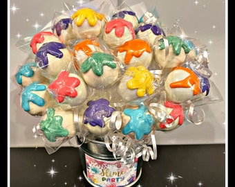 May include: A bouquet of cake pops decorated with colorful paint splatters, arranged in a metal bucket with a "Slime Party" label.