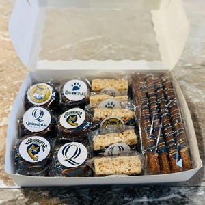 May include: A white box filled with assorted treats, including chocolate-covered cookies and pretzels, and rice crispy treats. The cookies have a Quinnipiac University logo. The pretzels are drizzled with blue and white icing.
