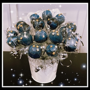 May include: A bouquet of blue and gold cake pops decorated with white and gold stars, arranged in a white bucket.