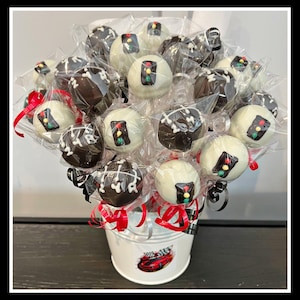May include: A white bucket with a red racing stripe and a red car graphic holds a bouquet of cake pops. The cake pops are decorated with black and white chocolate and traffic light designs.
