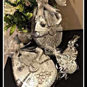 May include: Three silver Millennium Falcon spaceship-shaped favors wrapped in clear cellophane with silver ribbon bows. The favors are arranged on a black surface. The image includes the text "@sparklesbabyinc".