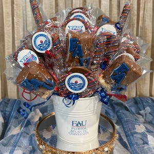 May include: A decorative arrangement of chocolate-covered treats in a white bucket. The treats include pretzels and cookies, decorated with blue, red, and white sprinkles and the letters "FAU". The bucket has the text "FAU FLORIDA ATLANTIC UNIVERSITY".