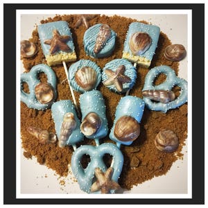 May include: An assortment of sea-themed treats, including blue-frosted pretzels, cookies, and rice crispy treats, decorated with chocolate seashells and starfish. The treats are arranged on a bed of brown crumbs, creating a beach-like scene.