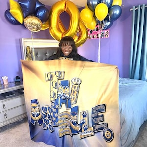May include: A tan and blue blanket with the text "TAI ARRELE" and "Quinnipiac" in a collegiate style. The blanket is held in front of a person. Gold and blue balloons are in the background, including balloons shaped like the numbers "80".