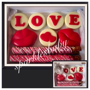 May include: A white gift box filled with chocolate treats. The box contains chocolate pretzels, chocolate lips, and chocolate circles with the word "LOVE" spelled out. The box is decorated with a pink and white ribbon.