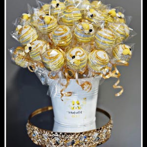 May include: A bouquet of bee-themed cake pops in a white bucket, presented on a gold-trimmed mirrored tray. The cake pops are yellow, gray, and gold, wrapped in clear cellophane. The bucket has a bee graphic and the text "What will it BEE?"