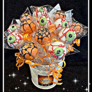 May include: A white metal bucket filled with a bouquet of Halloween-themed cake pops. The cake pops are decorated with orange, white, and black frosting and have googly eyes. The bucket has a "Happy Halloween" label.