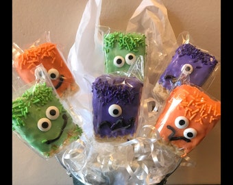 Puede incluir: Seis golosinas de arroz crujiente con forma de monstruo en palitos, decoradas con chispas y ojos saltones. Las golosinas están dispuestas en un cubo de metal con papel de seda blanco.