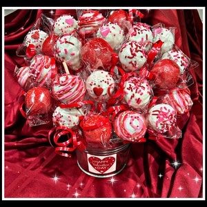 May include: A bouquet of red, white, and pink cake pops decorated with sprinkles and hearts. The cake pops are arranged in a metal bucket with a "Happy Valentine's Day" label. The bouquet is surrounded by red fabric.