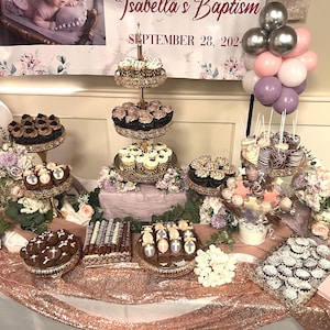 May include: A dessert table decorated with a pink and purple balloon arch, flowers, and various desserts, including cupcakes, cake pops, and chocolate-covered treats. The table is covered in a rose gold tablecloth. The sign reads "Isabella's Baptism September 28, 2022".