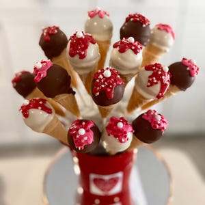 May include: A bouquet of cake pops decorated with white and pink frosting and sprinkles, arranged in a red vase with a heart design. The cake pops are on sticks and are in the shape of ice cream cones.