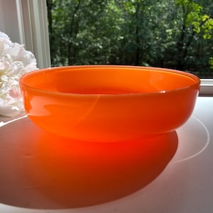 May include: An orange glass bowl with a rounded shape. The bowl is sitting on a white surface, with a white flower and green foliage in the background. The bowl casts a large shadow.