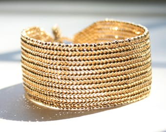 May include: A gold-toned metal bracelet with a woven chain design. The bracelet is made of multiple rows of small, linked metal pieces.