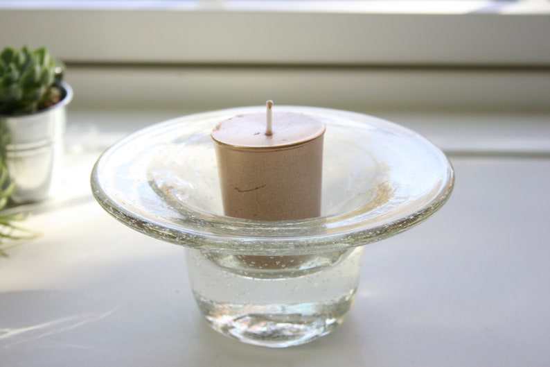 May include: A clear glass candle holder with a wide, flat base and a round, open top. A small, brown candle sits in the center of the holder.