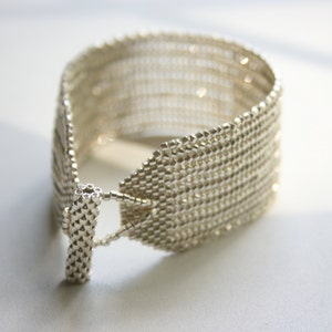 May include: A silver bracelet made of small, tightly woven beads. The bracelet is open on one side and has a clasp on the other.
