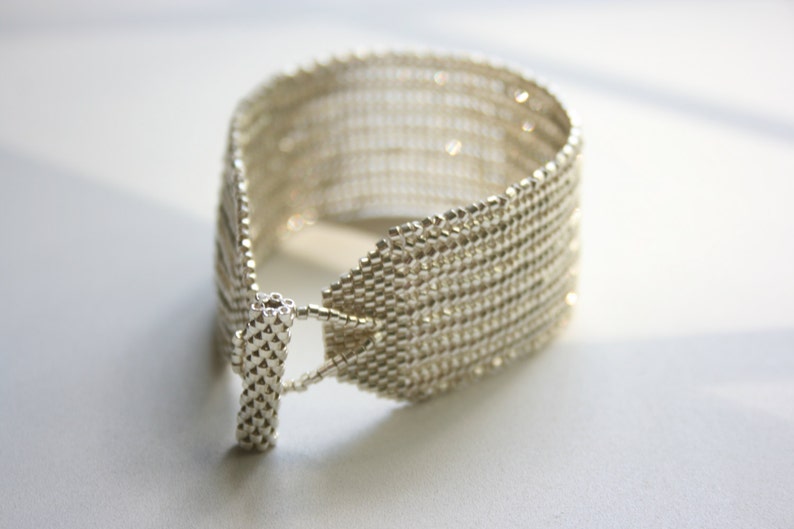 May include: A silver bracelet made of small, tightly woven beads. The bracelet is open on one side and has a clasp on the other.