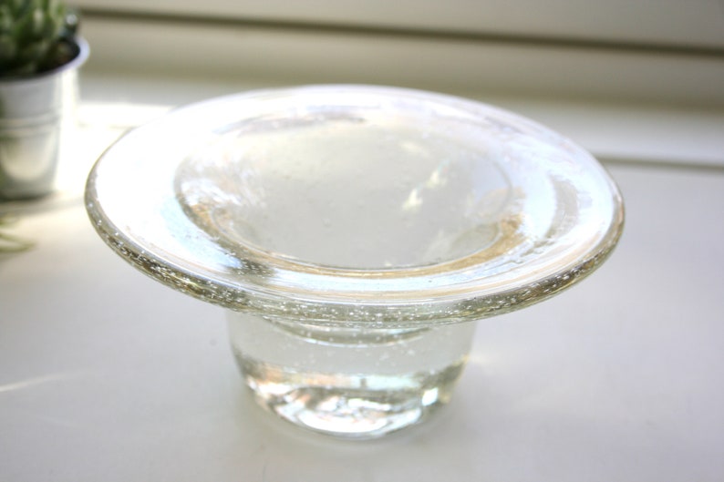 May include: A clear glass candle holder with a wide, flat top and a round base. The glass has a textured surface with small air bubbles.
