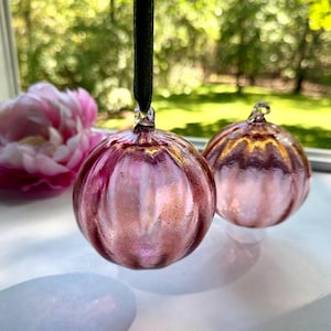 May include: Two round, ribbed glass ornaments in a translucent pink hue with a subtle metallic sheen. One ornament hangs from a dark green ribbon, while the other rests on a surface. Both feature a small glass loop at the top for hanging.