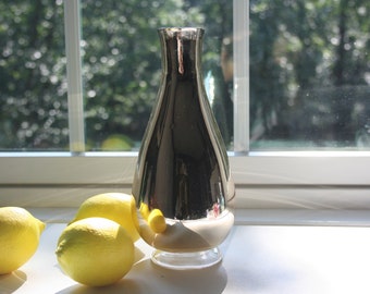 Puede incluir: Un jarrón de vidrio plateado y nacarado con forma redondeada de lágrima se encuentra sobre una superficie blanca. Tres limones amarillos están delante del jarrón.