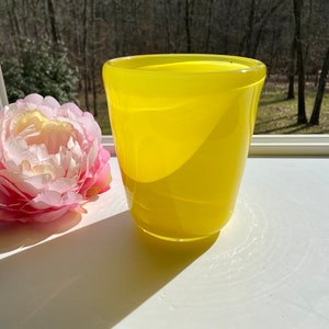 May include: A bright yellow glass vase with a rounded, slightly flared top. The vase is sitting on a white surface.