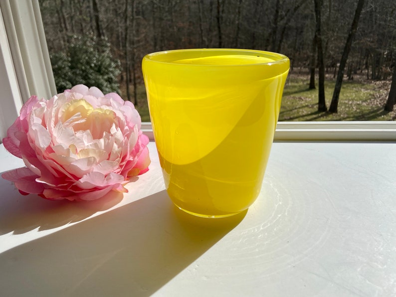 May include: A bright yellow glass vase with a rounded, slightly flared top. The vase is sitting on a white surface.
