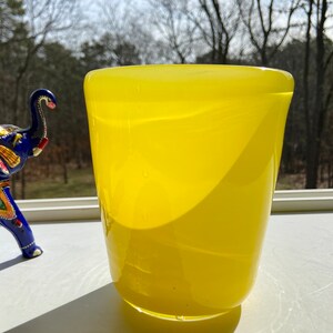 May include: A bright yellow glass vase with a rounded bottom and a wide opening. The vase is sitting on a white surface with a blurred background of trees.