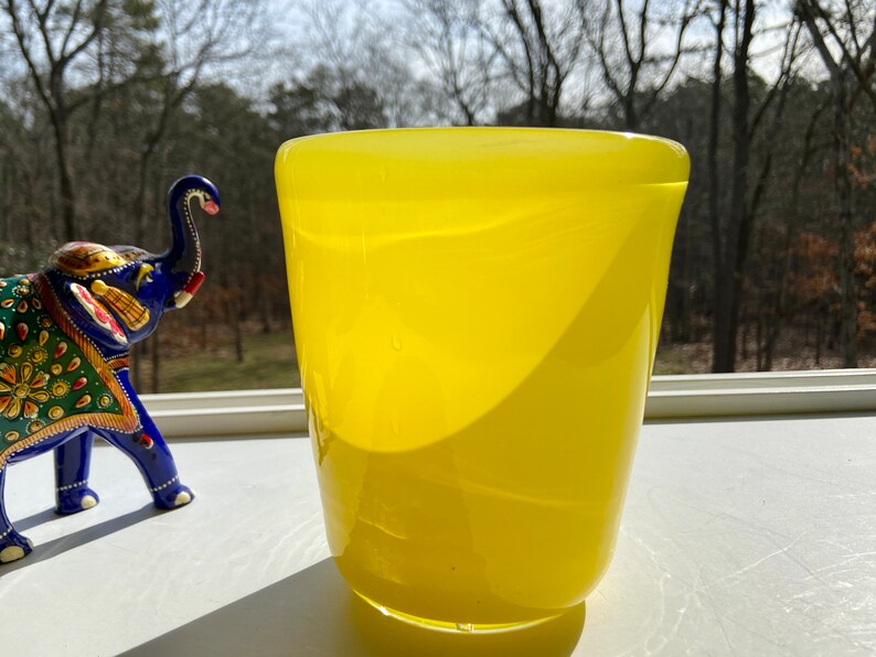 May include: A bright yellow glass vase with a rounded bottom and a wide opening. The vase is sitting on a white surface with a blurred background of trees.