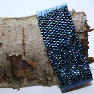 May include: A beaded bracelet with a shimmering, iridescent blue-green color. The bracelet is made of small, square beads arranged in a grid pattern. The bracelet is resting on a piece of textured, light brown bark.