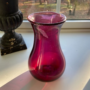 May include: A deep purple glass vase with a rounded bottom and a narrow neck. The vase is sitting on a white surface.