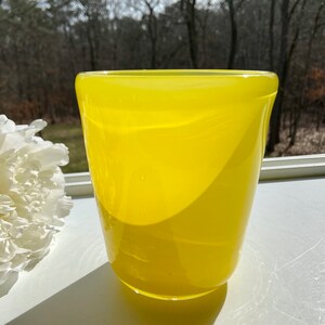 May include: A bright yellow glass vase with a rounded, slightly flared opening. The vase is sitting on a white surface.