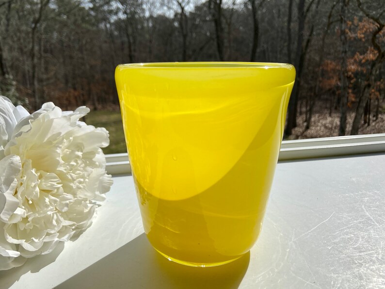 May include: A bright yellow glass vase with a rounded, slightly flared opening. The vase is sitting on a white surface.